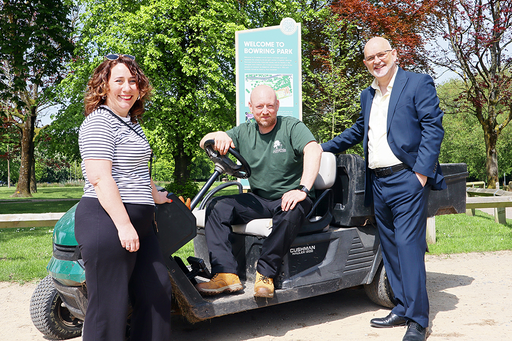 Golf greenskeeper sat on lawnmower with male and female representatives either side